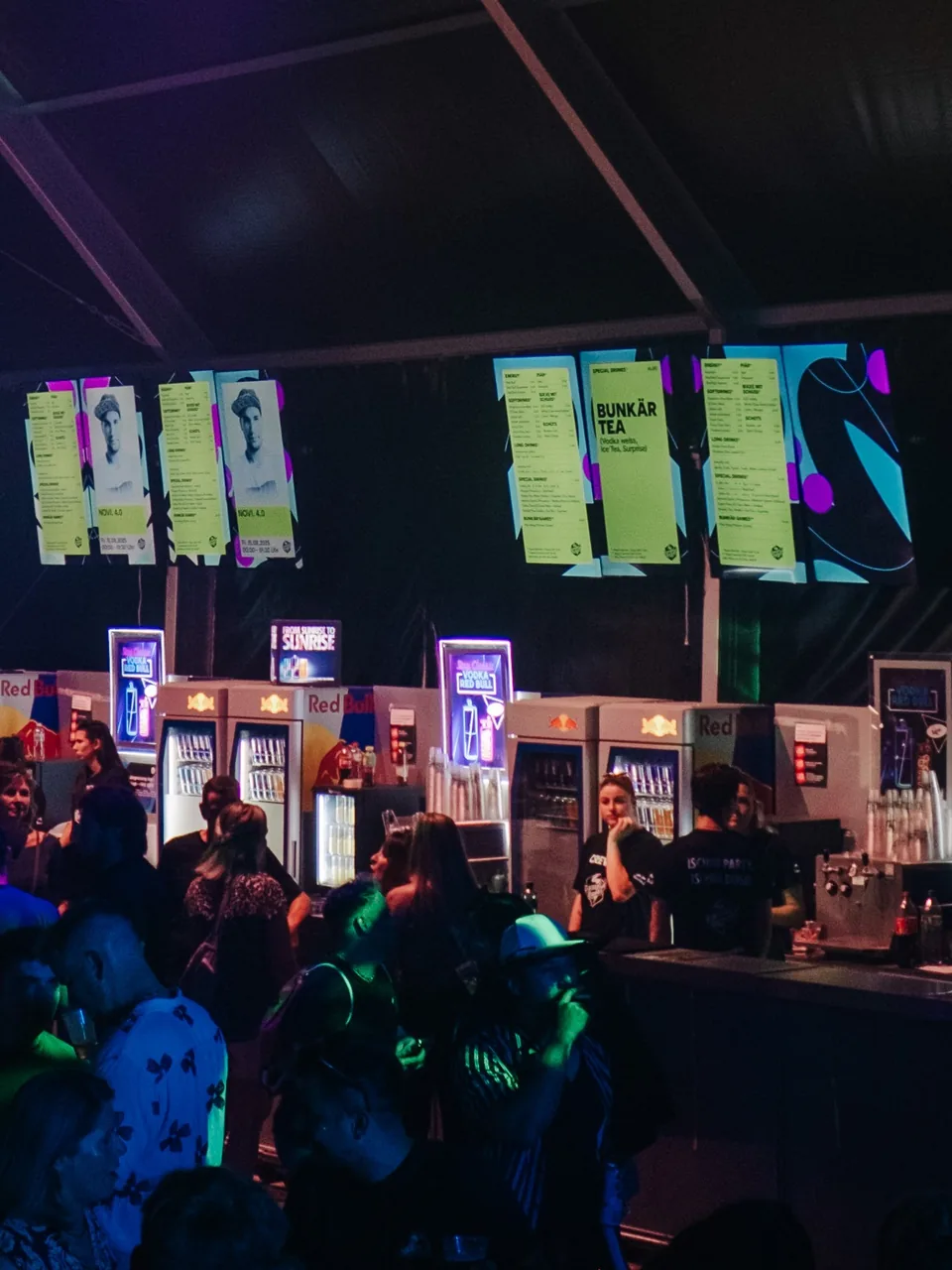 Bildschirme mit Getränkekarten über einer Festivalbar, davor Gäste.